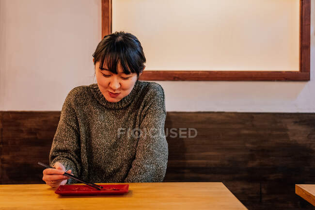 Молодая азиатка в свитере ест за деревянным столиком в кафе — стоковое фото