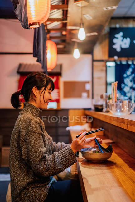 Vista lateral de la mujer asiática complacida en ropa casual riendo mientras se sienta en el mostrador con palillos y tazón con ramen en la cafetería - foto de stock