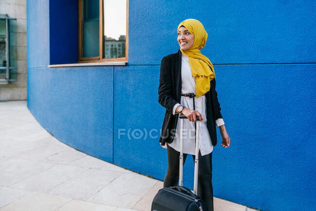Allegro musulmana femmina in velo in piedi con i bagagli in strada contro il muro blu — Foto stock