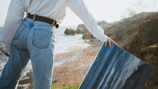 Возвращение молодой художницы в стильной одежде и шляпе, стоящей рядом с песчаным пляжем волнистого моря с живописью в руке — стоковое фото