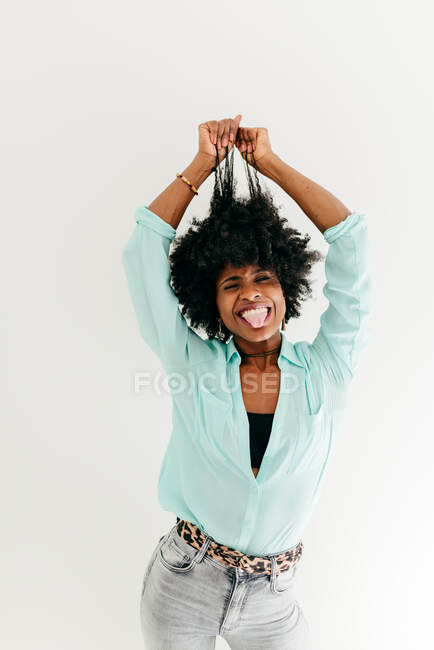 Playful young African American female in trendy outfit having fun touching afro hair on white background — Stock Photo