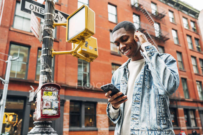 Desde abajo contenido alegre chico afroamericano en denim chaqueta de moda surfeando teléfono móvil moderno durante paseo por la ciudad - foto de stock