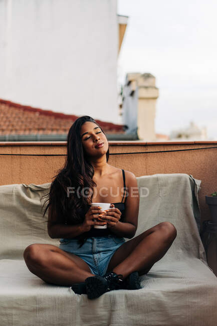 Junge hispanische Frau mit geschlossenen Augen sitzt im Schneidersitz auf dem Sofa und genießt Heißgetränk, während sie sich morgens auf dem Balkon ausruht — Stockfoto