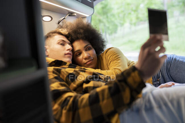 Diverse young man and woman looking on instant shot while chilling on bed in van during road trip in summer — Stock Photo