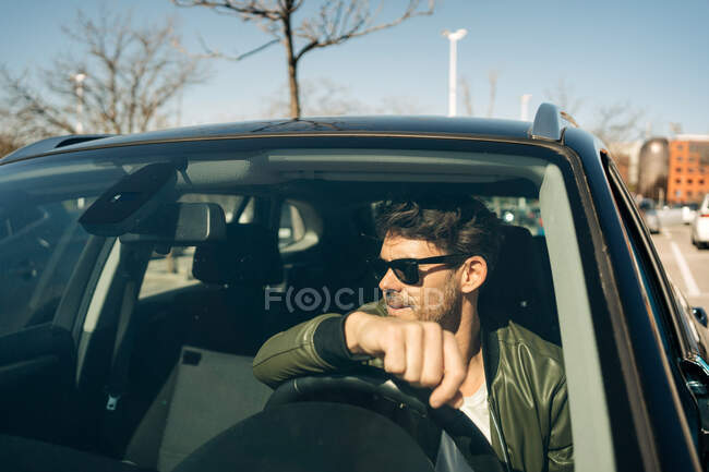 Brutal male in sunglasses driving modern automobile while looking away in city in sunlight — Stock Photo