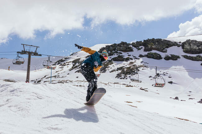 Atleta masculino anônimo em máscara de tecido pulando com snowboard sobre a neve contra a Serra Nevada e via cabo na Espanha — Fotografia de Stock