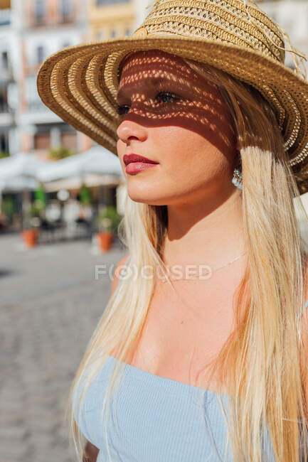Vue latérale de charmante femme portant un chapeau de paille regardant loin par une journée ensoleillée dans la rue de la ville en été — Photo de stock