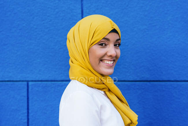 Vista lateral da fêmea muçulmana na moda em hijab amarelo em pé olhando para a câmera na rua no fundo azul — Fotografia de Stock