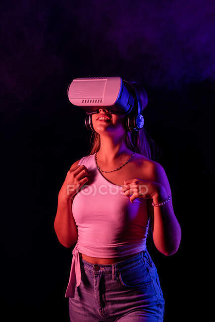 Unerkennbare Frau im VR-Headset, die mit virtueller Realität interagiert, während sie im dunklen Studio mit Dampf und blauer und pinkfarbener Neonbeleuchtung steht — Stockfoto