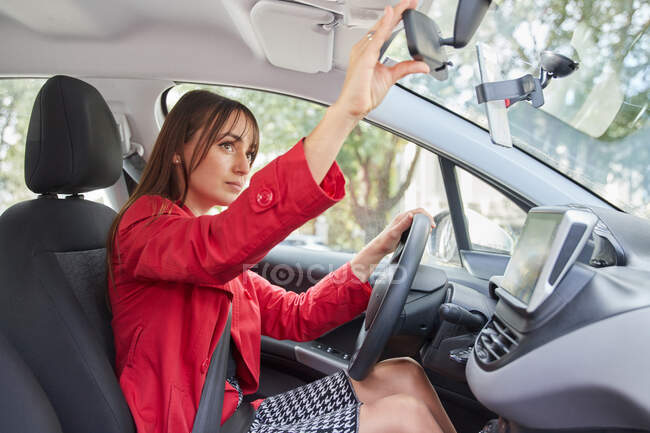Vista lateral de la joven conductora en chaqueta roja de moda que prepara el espejo retrovisor para el viaje dentro del automóvil moderno - foto de stock
