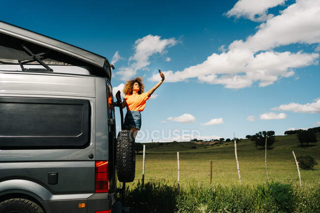 Femme noire avec les cheveux bouclés debout sur l'échelle van extérieur et à la recherche d'une couverture pendant le voyage sur la route — Photo de stock