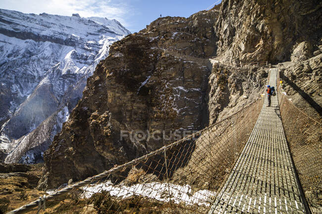 Vista lateral do mochileiro irreconhecível em pé na ponte suspensa de metal e admirando a vista das montanhas rochosas do Himalaia no dia ensolarado no Nepal — Fotografia de Stock