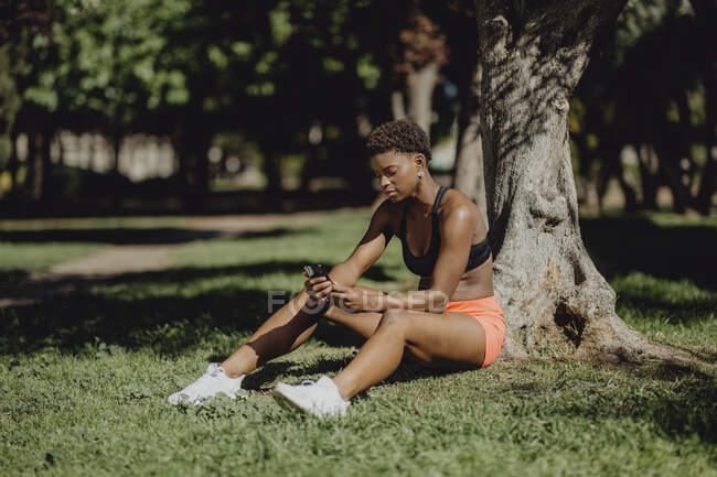 African American woman in sportswear holding smartphone while sitting on grass in sunny day — Stock Photo