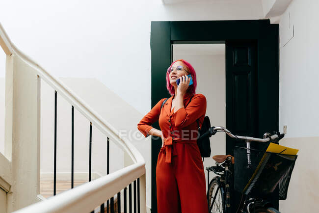 Jeune femme élégante en costume rouge avec sac à dos parlant sur smartphone tout en se tenant debout avec vélo sur escalier — Photo de stock