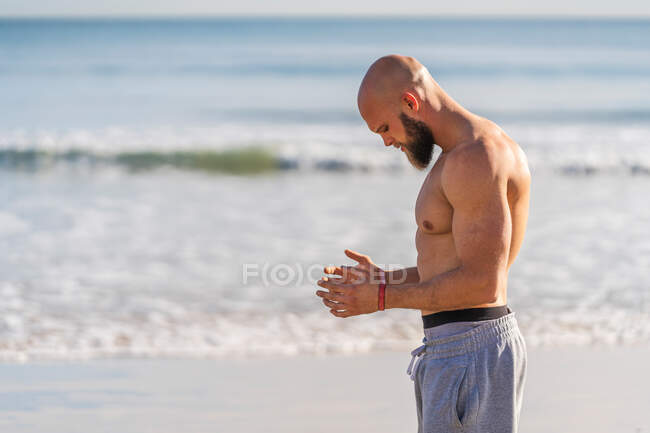 Vista lateral de atleta masculino sin camisa con banda elástica mirando hacia abajo mientras hace ejercicio en la playa soleada vacía - foto de stock
