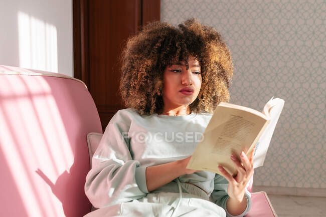 Зосереджена афроамериканка сидить вдома на дивані і читає цікаву книжку. — стокове фото
