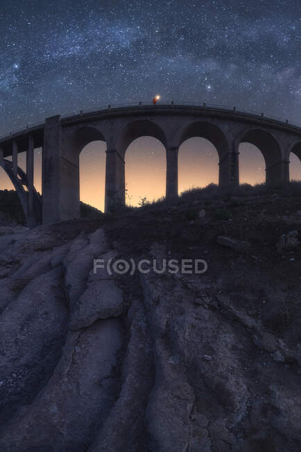 Increíble paisaje de envejecido puente de piedra con elementos arqueados que cruzan el río bajo el cielo nocturno con la brillante Vía Láctea y la luz del atardecer - foto de stock