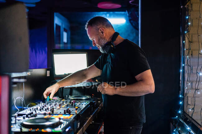 Side view pensive middle aged man in headphones and black casual clothes focusing and playing professional DJ controller while performing at concert in contemporary night club against laptop with blank screen — Stock Photo
