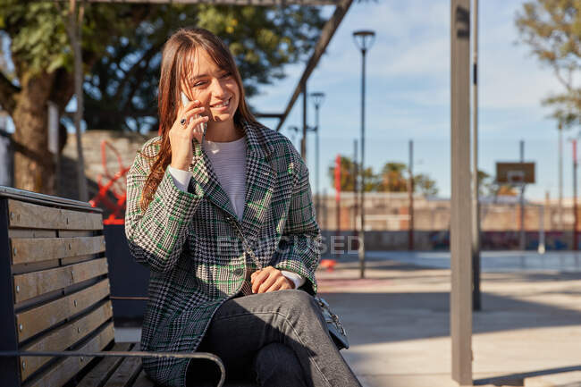 Moderna hembra milenaria sonriente con elegante atuendo de primavera sentada en el banco y respondiendo a una llamada telefónica mientras descansa en la calle urbana mirando hacia otro lado - foto de stock