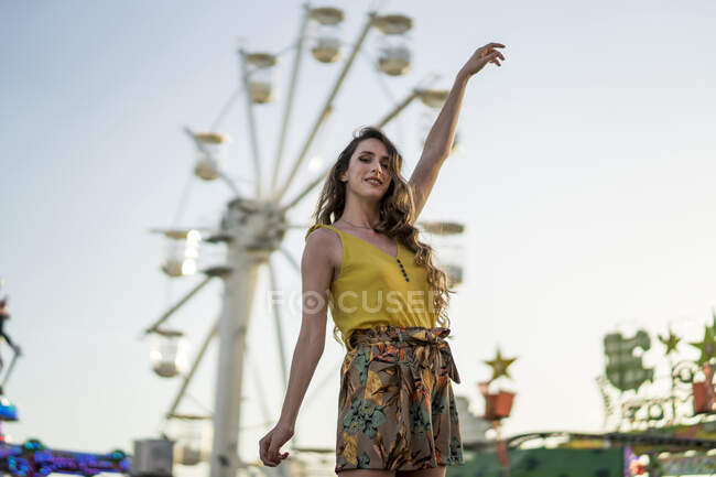 Baixo ângulo de mulher encantada em pé olhando para a câmera com braços levantados no parque de diversões e desfrutando de fim de semana de verão — Fotografia de Stock