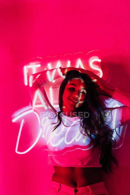 Young Hispanic female touching hair and looking at camera while leaning on wall near neon It Was All Dream inscription — Stock Photo