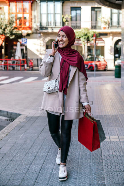 Веселая мусульманка в хиджабе и с пакетами для покупок ходит по улице и разговаривает по смартфону — стоковое фото