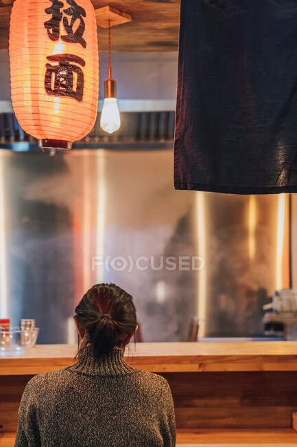 Vista posteriore della donna dai capelli neri senza volto in maglione seduto al bancone in accogliente bar ramen — Foto stock