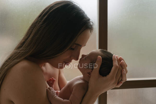 Vista lateral da mãe macia com os olhos fechados de pé com bebê nu bonito perto da janela em casa — Fotografia de Stock