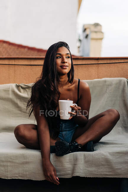 Joven mujer hispana sentada con las piernas cruzadas en el sofá mirando hacia otro lado y disfrutando de una bebida caliente mientras descansa en el balcón por la mañana - foto de stock