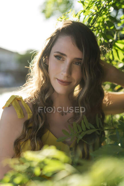 Serena fêmea com cabelo ondulado em pé perto de árvore verde no jardim no verão e olhando para longe — Fotografia de Stock