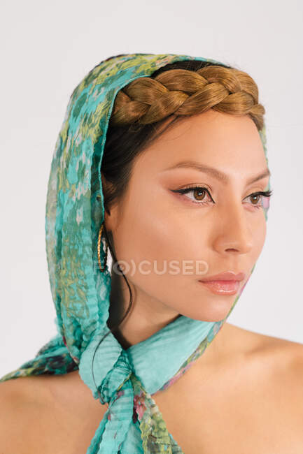 Modelo femenino joven con peinado trenzado y maquillaje elegante con pañuelo colorido con estampado floral verde mirando hacia otro lado sobre fondo blanco - foto de stock