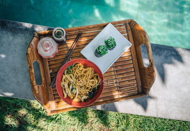 Vista superior de la bandeja con rollos de sushi gunkan con chuka contra un tazón de pasta cocida y trozos de pollo junto a la piscina - foto de stock