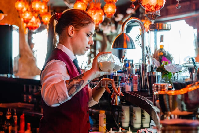 Garçonete feminina derramando álcool em shaker enquanto prepara coquetel refrescante no balcão no bar e olhando para longe — Fotografia de Stock