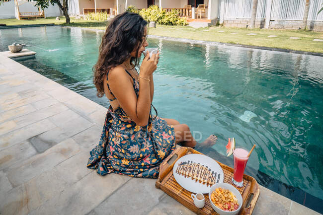 Viajero femenino con vista lateral con bebida caliente mirando hacia otro lado mientras está sentado en la piscina con sabroso desayuno en bandeja - foto de stock