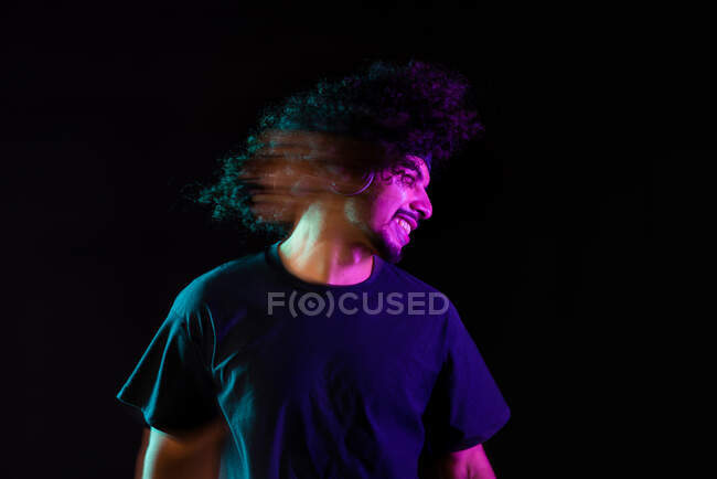 Delizioso maschio latino in cuffia che ascolta musica e getta capelli ricci in studio con luci al neon su sfondo nero — Foto stock