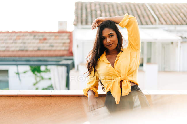 Молодая латиноамериканка в повседневной одежде, опираясь на перила и глядя в камеру, днем отдыхает на балконе — стоковое фото