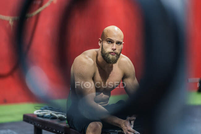 Vista lateral do homem musculoso exausto olhando para a câmera sentada em pesos e descansando durante o treino funcional no ginásio — Fotografia de Stock