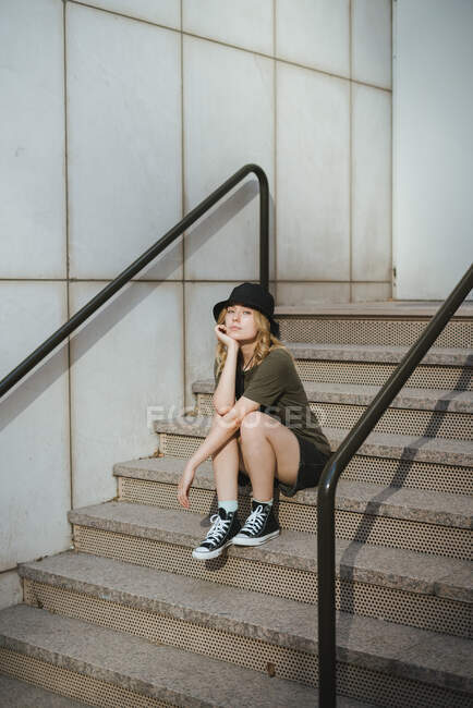 Молодая женщина в повседневной одежде смотрит на камеру, сидящую на лестнице напротив бетонной стены современного здания на городской улице днем — стоковое фото