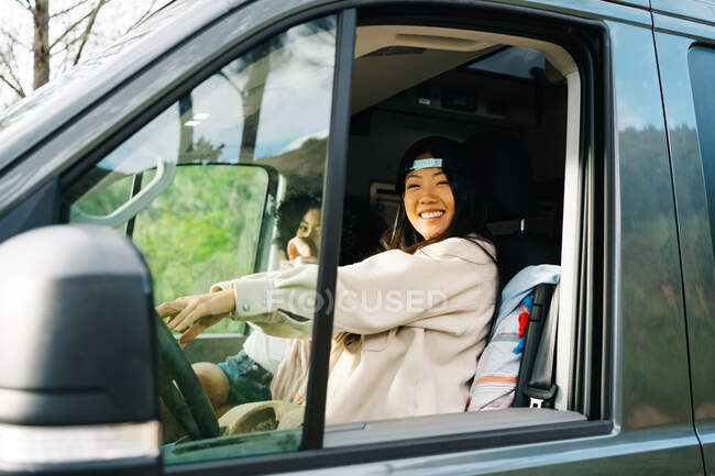 Through window side view of cheerful young Asian woman sitting on driver seat of camper vehicle and looking at camera while traveling with girlfriend through nature — Stock Photo