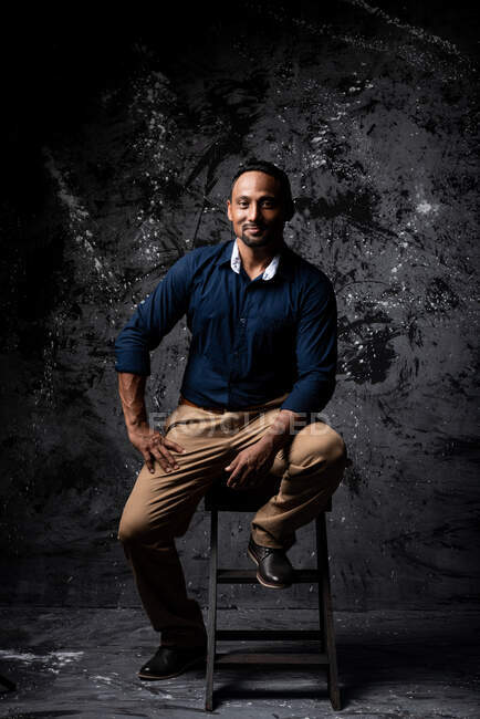 Delighted stylish ethnic male sitting on wooden stool in studio on dark background and looking at camera — Stock Photo