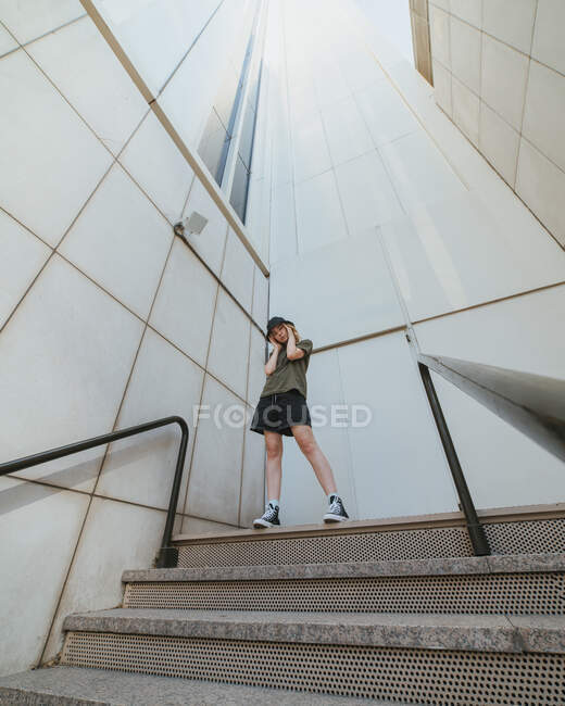 From below of young female in casual apparel touching face on city staircase against multistory house — Stock Photo