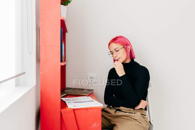 Jovem designer feminina criativa pensativa com cabelo rosa em roupas casuais e óculos desenhando esboço enquanto trabalhava na mesa em casa — Fotografia de Stock