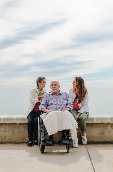 Happy family having fun with aged father sitting in wheelchair on embankment near sea on sunny day — Stock Photo