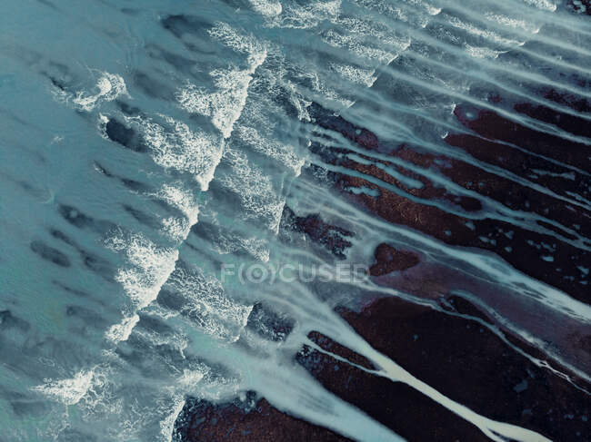 Desde arriba paisaje texturizado aéreo de áspera costa pedregosa desigual y espumosas olas oceánicas con arroyos - foto de stock