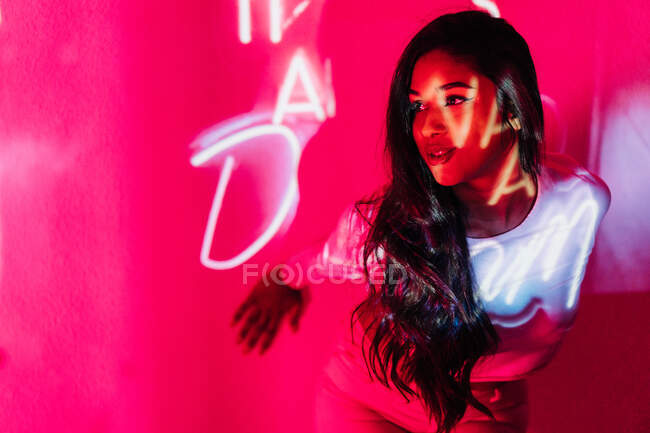 Junge hispanische Frau schaut weg, während sie sich an Wand in der Nähe von Neon lehnt — Stockfoto