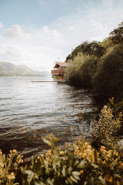 Maison confortable avec jetée située sur la côte du lac ondulant contre le ciel nuageux dans la campagne britannique — Photo de stock