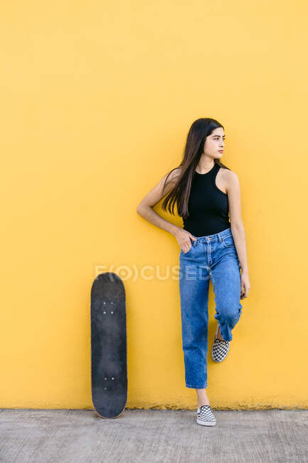 Jeune patineuse avec planche à roulettes debout regardant loin sur la passerelle avec mur jaune coloré sur le fond en journée — Photo de stock