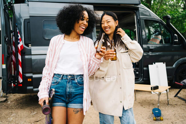 Fröhliche junge multiethnische Freundinnen, die Flaschen Bier klappern, während sie Spaß haben und gemeinsam die Sommerferien in der Nähe des in der Natur geparkten Wohnmobils genießen — Stockfoto