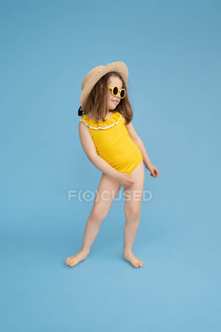 Corpo pieno di simpatica bambina felice indossando costume da bagno giallo e cappello di paglia con eleganti occhiali da sole in piedi su sfondo blu e guardando altrove — Foto stock