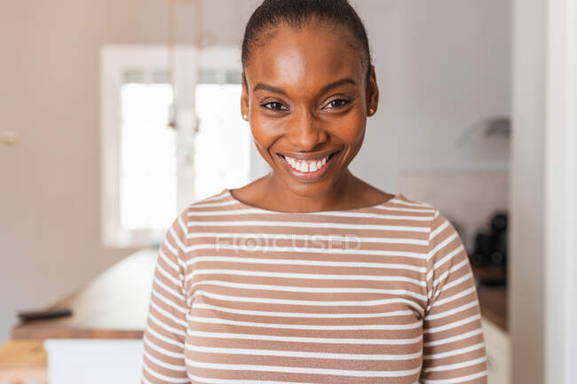 Feliz joven afroamericana hembra con moño de pelo mirando a la cámara contra la mesa y lavadora en la cocina - foto de stock